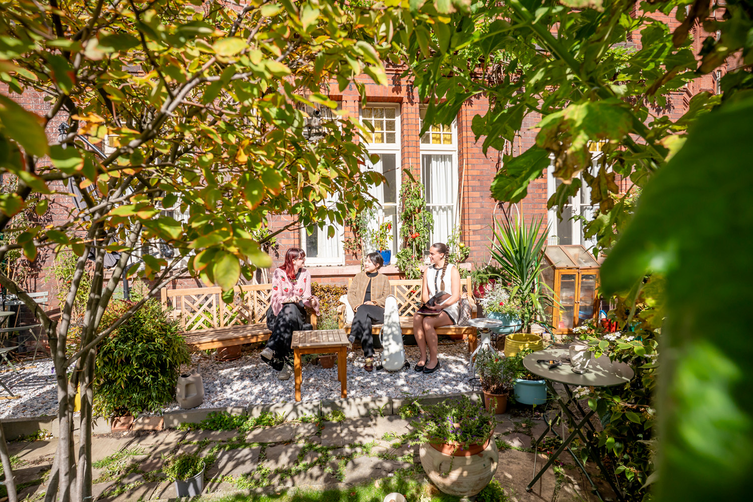 Students sitting on a wooden bench in a small secluded garden with a red brick wall behind them.